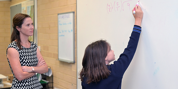 Teacher watching student doing sums on whiteboard.