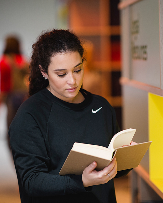 Female student standing in library reviewing a book
